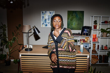 Portrait of young adult Black man wearing glasses standing in modern workspace touching chin thoughtfully, second hand context indicated by eclectic decor and reused furnishings