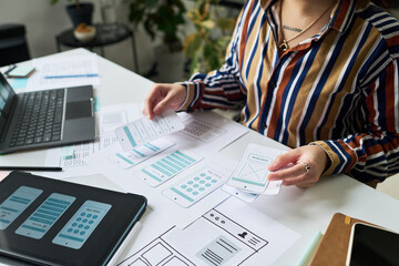 Anonymous woman working on mobile app interface design, arranging paper wireframes and prototypes on desk with laptop and tablet, focusing on user experience planning