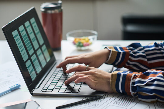 Unknown woman working on laptop, designing mobile application interface, hands typing on keyboard, digital wireframes visible on screen, office desk in background
