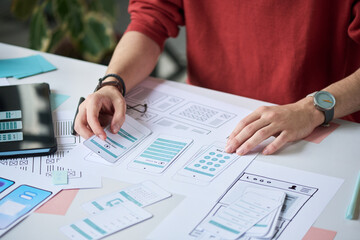 Unknown man working on mobile app interface design, arranging paper prototypes and wireframes on desk, using hands to organize layout and plan user experience