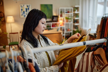 Asian young adult woman browsing second hand clothing rack, examining garment closely in thrift store setting, surrounded by various clothes, focused on sustainable shopping