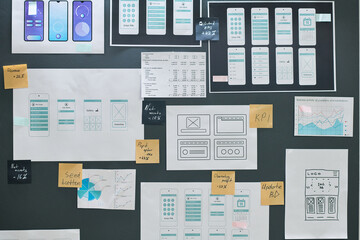 Mobile app wireframes and user interface sketches arranged on wall with printed charts, graphs, and sticky notes showing project planning, design process, and workflow organization