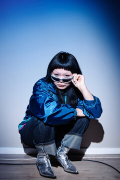Asian young adult woman crouching near wall, holding sunglasses and looking directly at camera, wearing metallic boots, embodying second hand fashion style in urban setting