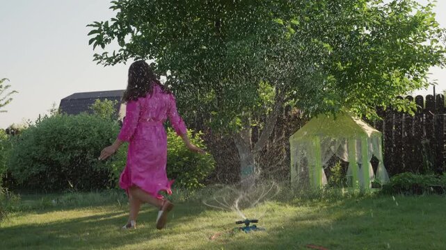 Woman In Bright Pink Dress Running Through Sprinkler In Sunny Garden, Water Spray And Energetic Movement Create Playful Summer Atmosphere With Backyard Greenery And Carefree Motion