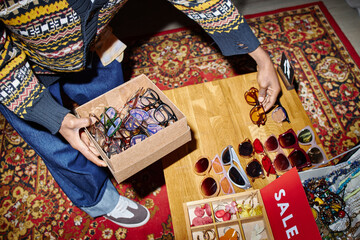 Young adult Black man sorting second hand sunglasses on wooden table, holding cardboard box filled...