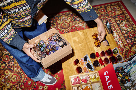Young adult Black man sorting second hand sunglasses on wooden table, holding cardboard box filled with assorted eyewear, arranging items for sale in thrift shop setting