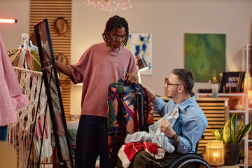 Black young adult man showing patterned shirt to Caucasian middle aged man with disability in wheelchair, shopping for second hand clothing in modern store interior