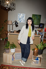 Young adult Asian woman standing in modern workspace leaning against wooden shelf with second hand sign visible, surrounded by potted plants and creative supplies