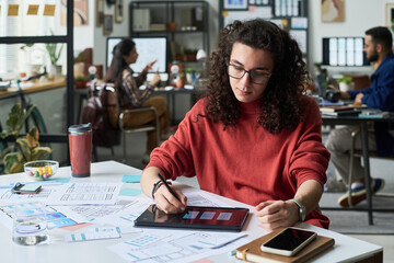 Young adult Caucasian man using digital tablet stylus while working on user interface design sketches at desk in modern office, colleagues collaborating in background, smartphone and coffee cup nearby