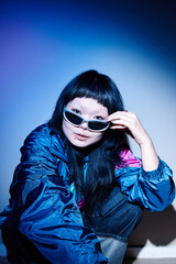 Portrait of young adult Asian woman sitting on floor adjusting sunglasses with confident expression, wearing second hand clothing, looking directly at camera in studio setting