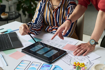 Unknown woman collaborating with man using digital tablet for mobile app interface design, hands pointing at wireframe sketches on desk