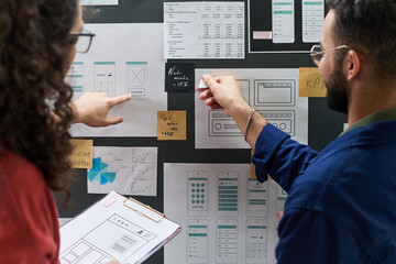 Two coworkers collaborating on user interface design project, discussing wireframes and data charts on wall, both pointing at printed layouts and notes