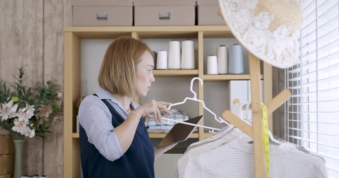Asian adult female dressmaker using digital tablet to review fashion design in workspace surrounded by textile, sewing tools and clothing racks inside small fashion business studio