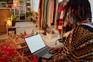 Young adult Black woman browsing online marketplace using laptop in second hand clothing store, sitting near racks of pre owned garments and accessories, focused on screen