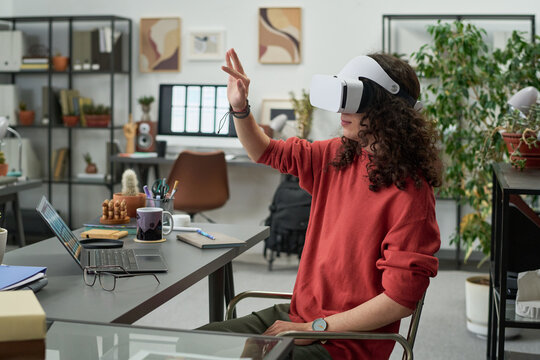 Young adult Caucasian man wearing virtual reality headset interacting with digital environment, while sitting at desk in modern office workspace with laptop and plants visible - Powered by Adobe
