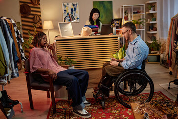 Fototapeta premium Black young adult man sitting and talking with Caucasian middle aged man with disability in wheelchair in second hand shop, while Caucasian young adult woman working at counter in background