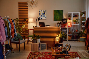 Second hand clothing store interior showing wooden counter with laptop, racks of assorted garments, decorative plants, shelving with accessories, framed sign reading second hand