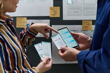 Young woman and man collaborating on mobile app interface design, holding smartphone and wireframe cards, discussing user experience in office setting