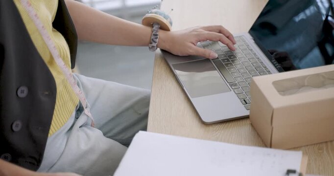 closeup hands young asian female designer sketching fashion idea using laptop digital pen drawing garment pattern workspace filled fabric sewing tools natural tone emphasizing creativity process
