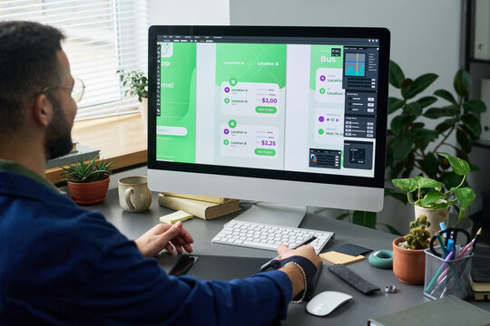 Man working on user interface design for mobile application on desktop computer, sitting at desk with digital pen and looking at large monitor displaying app screens - Powered by Adobe