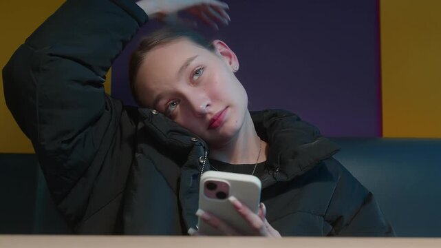 Teen girl in black jacket crunches the cervical vertebrae while sitting at cafe table holding smartphone, indoor evening light, symbolizing fast food lifestyle and mobile communication.
