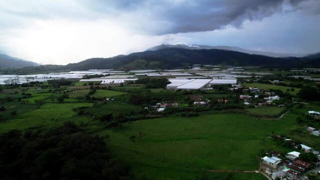 Ascending drone shot revealing greenhouse farming facilities surrounded by green valleys and mountains in Jarabacoa under dramatic cloudy skies.