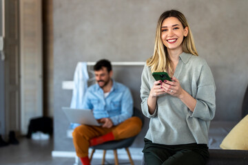Smiling caucasian woman using her smartphone while her partner works in the background, home office