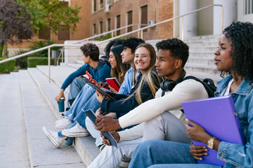 Diverse group of college students sitting on campus stairs studying, chatting and using devices together, smiling and collaborative