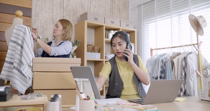 Asian female fashion designer placing new fabric sample on mannequin while young asian assistant working nearby inside tailoring studio preparing clothing products for delivery and ecommerce order