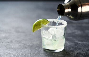 Margarita cocktail is poured from a shaker into a glass with a slice of lime and an edge of salt along the edge