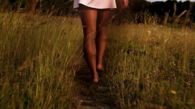 black woman in skirt walks barefoot through path in field with high grass during a warm summer day.