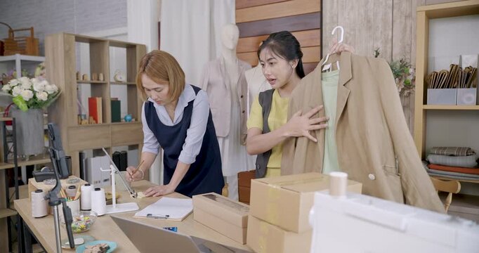 Asian female fashion designer presenting handmade jacket beside coworker preparing parcels for shipping in modern tailoring studio representing small fashion business environment