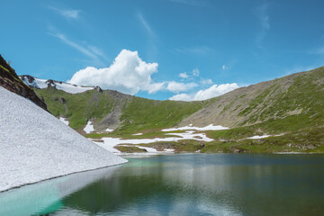 Scenic view to beautiful glacial lake in mountain cirque. Sunlit scenery with turquoise alpine lake surrounded by white ice and green rocky ridge under lush clouds in blue sky. High mountain lake.