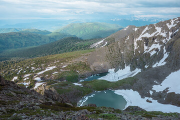 Awesome harsh top view from rock precipice edge to two turquoise alpine lakes with ice floes in stone mountain cirque. Small creek between two green icy lakes in rocky stony terrain under cloudy sky.