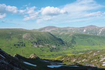 Scenic upland hilly view above sheer crags along rock ridge to green alpine valley. Steep rocky slope with pointy peaks against forest on big hills under clouds in blue sky. Aerial top view from above