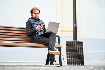 Man sits on wooden bench, working on laptop and smartphone, both connected to solar panel. Urban setting and casual attire highlight modern, sustainable, mobile workspace.