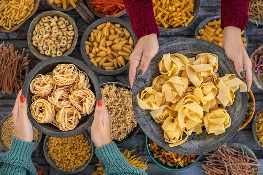Different types of uncooked pasta at the hands of two women in the wood bowls.