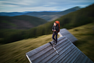 Hiker with orange backpack stands on roof of wooden cabin, surrounded by blurred, dynamic scenery. Motion blur effect.