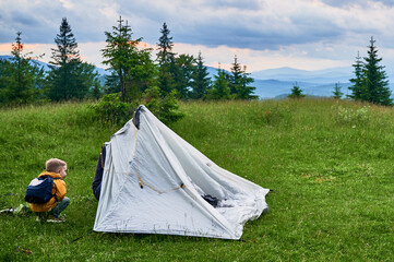 Young child with backpack standing near white tent being set up in scenic meadow. Pine trees and distant mountains under cloudy sky, creating serene outdoor camping scene.