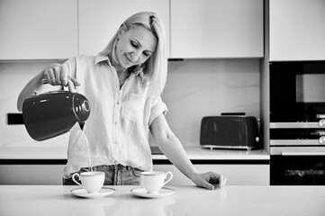 Young woman making morning coffee. Pretty female filling cup with hot water. Woman holding kettle and pouring water into cup. Black and white image.