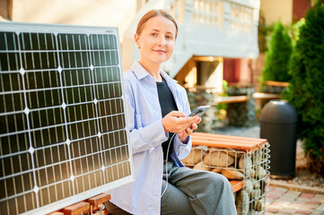 Happy woman using smartphone connected to photovoltaic solar panel. Integration of sustainable renewable energy into everyday life, demonstrating practical use of solar power for charging devices.
