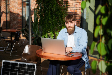 Man works on laptop and smartphone at outdoor table. Solar panel nearby, emphasizing sustainable, modern eco-friendly workspace that combines technology and sustainability.