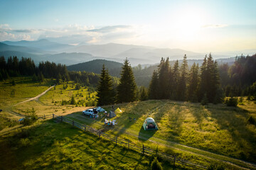 Aerial view of picturesque campsite on grassy hill, surrounded by evergreen trees and overlooking mountain vista. Distant mountains, tourist tents, cars, camping gear under golden evening light.