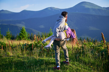 Father joyfully playing with his daughter, as they share playful moment in meadow. Both laughing, with backdrop of lush green hills and distant mountains under clear blue sky.