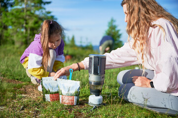 Tourist woman and young girl preparing meal from packaged pouch using portable camping stove on grassy hillside. Scenic mountain backdrop adds to serene outdoor setting.