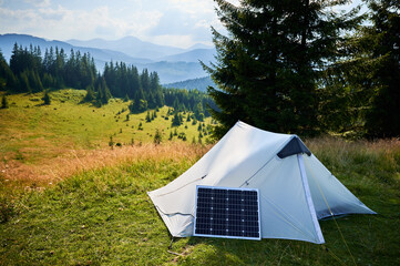 White tent equipped with solar panel sits in grassy field surrounded by lush trees and hills. Solar panel highlights eco-friendly approach to camping in tranquil, serene natural setting.