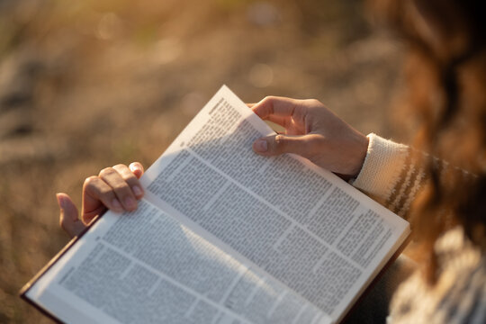 The image depicts a woman reading the Bible outdoors in soft sunlight. The concept encompasses reading the Bible and seeking God's words in the sacred texts, spiritual growth, and daily devotion.