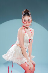 Elegant Asian model in white lace corset top and ruffled mini skirt with red ribbon accents, posing confidently in sheer red stockings and nude high heels, glamorous studio portrait soft blue scene