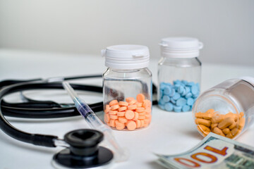 Assortment of prescription medications in clear vials next to a black stethoscope and syringe with currency.