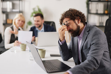 A group of workers struggles with business challenges in a modern office. One person shows signs of fatigue and stress while using a laptop. Others discuss the issues in the background.